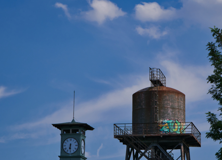 Very old partly rusty water tower with a clock tower in the foreground and blue sky in the...