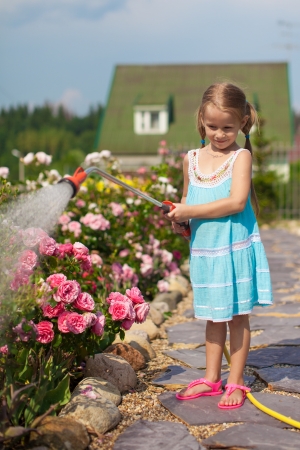Little girl in blue dress watering flowers with a hose in her gardenの写真素材