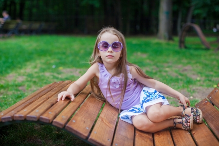 Cute little girl in purple glasses lying on wooden chair outdoorの写真素材