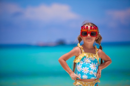 Adorable little girl in Happy Birthday glasses smiling and having fun at white beachの写真素材