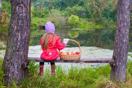 Rear view of a young girl in a jacket sits on a bench and holding a large basket with red applesの写真素材