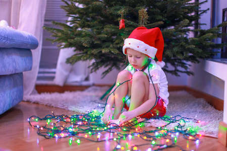 Little beautiful girl in Santa Claus hat sitting under the Christmas tree among garlandsの写真素材
