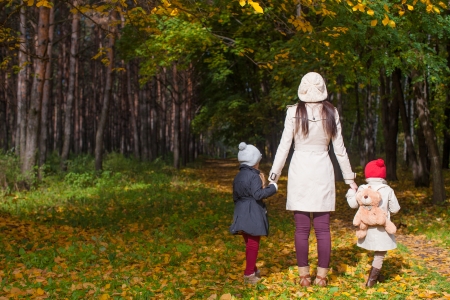Back view of young mother and her cute little daughters walking in autumn park at sunny dayの写真素材