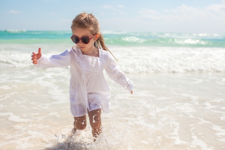Cute toddler girl in white dress standing in shallow water at exotic beachの写真素材