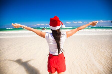 Back view of beautiful girl in santa hat walking like a bird on white beachの写真素材