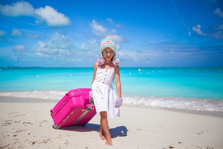 Adorable girl in warm winter hat and mittens walking with luggage on beachの写真素材