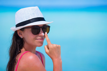Young happy woman applying suntan lotion on her nose on white beachの写真素材