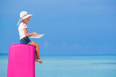 Little adorable girl with big pink suitcase and map on white beachの写真素材