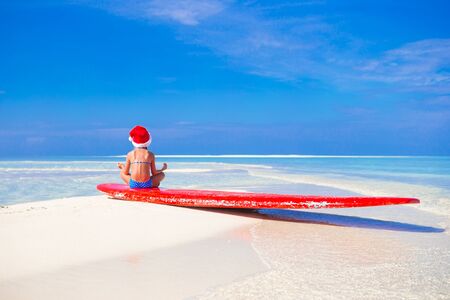 Adorable little girl in red Santa hat on surfboard during tropical vacation