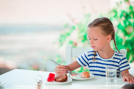 Adorable little girl having breakfast at cafe with sea viewの写真素材