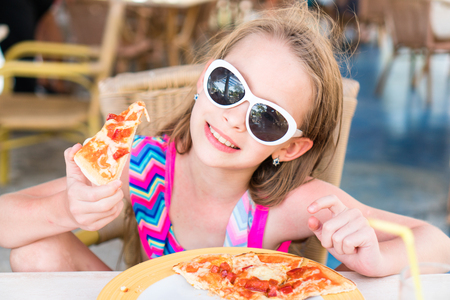 Portrait of cute little girl sitting by dinner table and eating pizzaの写真素材