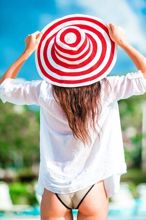 Beautiful young woman relaxing on the edge of infiniti swimming pool. Back view of girl in bikini and big red hatの写真素材