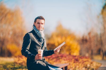 Young man drinking coffee with phone in autumn park outdoorsの写真素材
