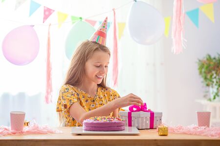 Caucasian girl is dreamily smiling and looking at birthday rainbow cake. Festive colorful background with balloons. Birthday party and wishes concept.の写真素材