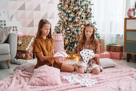 Adorable little girls sitting near the tree and making paper snow-flakes. Room decorated.の写真素材