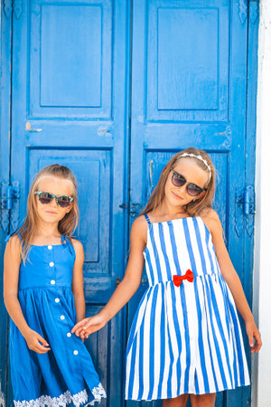 Little happy girls in dresses at street of typical greek traditional village on Mykonos Island, in...