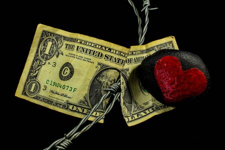 Barbed wire running through a old weathered and ripped dollar bill and a black stone with a painted red heart laying on itの写真素材