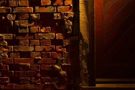 Old grungy and damaged red brick wall at night beside a wooden door with light of a lantern shining on itの写真素材