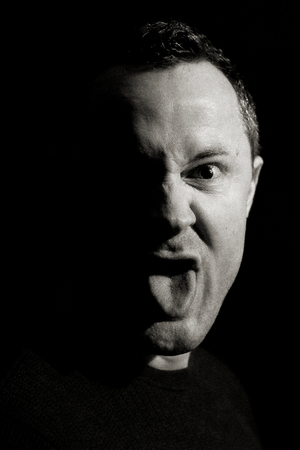 Stick out your tongue. Portrait of a man head between 30 and 40 years. His face shows the tongue in black and white soft Low Key Design and the view goes directly into the camera, the eye is wide open and the Face is angry or funnyの写真素材