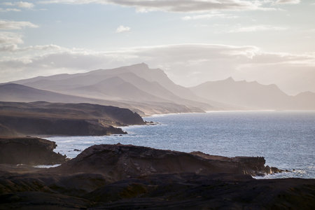 The rocky and indented west coast of Fuerteventura with the typical mountains in the light fog as...