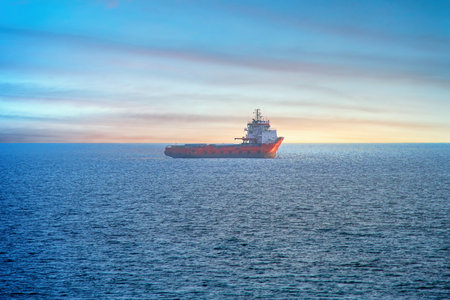 red cargo ship cruising on the cold open sea towards sunrise on the horizon, at the blue hour