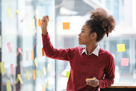 African American female employee write down on colorful sticky notes manage list, concentrated biracial woman work on startup brainstorm collaborate plan on stickers on glass wallの写真素材