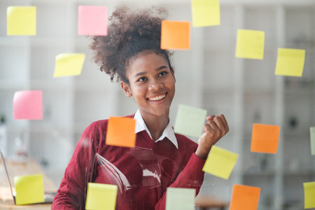 African American female employee write down on colorful sticky notes manage list, concentrated...