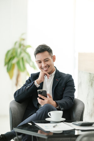 Successful and happy young Asian man, businessman, happy, shows with hand yes. Made a contract. Won money. Sitting with a smart phone or notebook at the table in a modern office.の写真素材