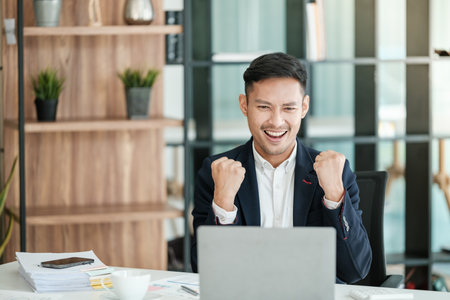 Successful and happy young Asian man, businessman, happy, shows with hand yes. Made a contract. Won money. Sitting with a notebook at the table in a modern office.の写真素材
