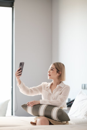 Cheerful beautiful Asian woman selfie or video call with smart phone on the bed.の写真素材