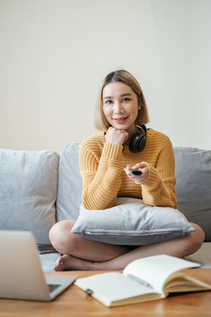 Selective focus on remote of Portrait beautiful young asian woman use remote on television in living room interior.の写真素材
