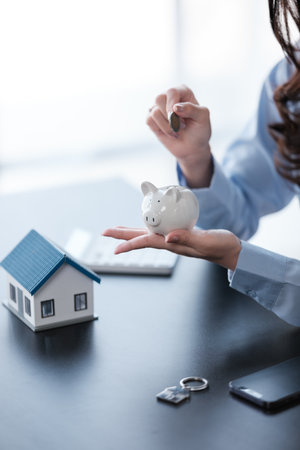 Close up Asian womens hand saving a coin into piggy bank with wooden house model on the table for business, finance, saving money and property investment concept.の写真素材