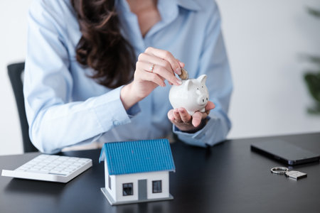 Close up Asian womens hand saving a coin into piggy bank with wooden house model on the table for business, finance, saving money and property investment concept.の写真素材