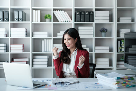 Cheerful Beautiful asian businesswoman working on laptop and calculating with financial calculator from statistics data graphs, charts. Successful business results in modern office wear red shirt.