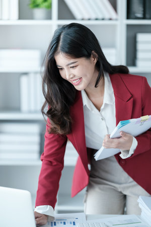 Beautiful asian businesswoman working on laptop and calculating with financial calculator from statistics data graphs, charts. Successful business results in modern office wear red shirt.の写真素材