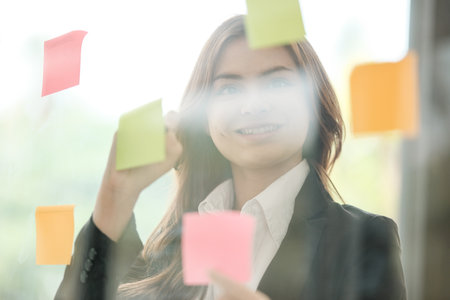Beautiful female employee write down on colorful sticky notes manage list, concentrated biracial woman work on startup brainstorm collaborate plan on stickers on glass wall.の写真素材
