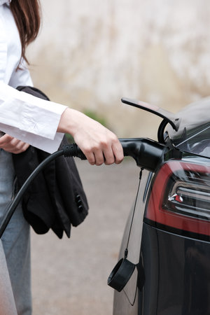 Young woman traveling by electric car having stop at charging station standing plugging charger close-up.の写真素材