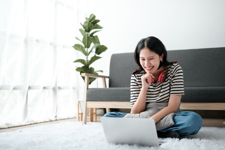Smiling young asian teen girl video calling on laptop. Happy mixed race pretty woman student looking at computer screen watching webinar or doing video chat by webcam.の写真素材