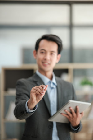 Smart and professional millennial Asian businessman in a formal business suit stands, isolated background with his portable tablet and pointing pen at camera.の写真素材