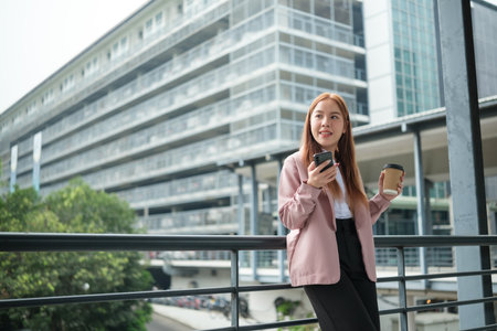 A young Asian businesswoman wearing a suit holding a cup of coffee in a big city. Young Asian businesswomen using a smart phone contact the clients.の写真素材