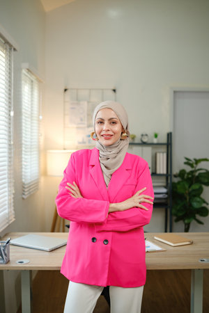 A woman in a pink jacket and white pants stands in front of a desk with a laptopの写真素材