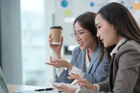 Two women are sitting at a desk, one holding a cup of coffeeの写真素材