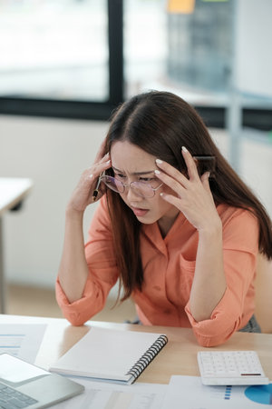 A woman in an orange shirt is sitting at a desk with a notebook and a calculatorの写真素材