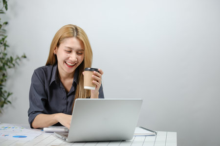 A woman is sitting at a desk with a laptop and a cup of coffeeの写真素材