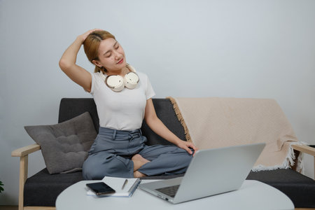 A woman is sitting on a couch with her head tilted and a laptop in front of herの写真素材