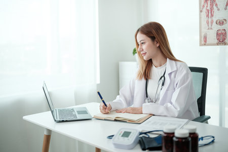 A woman doctor is sitting at a desk with a laptop and a notebookの写真素材