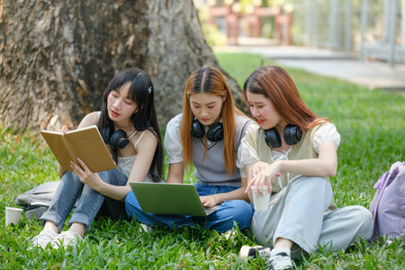 Three young women are sitting on the grass, reading books and using a laptopの写真素材