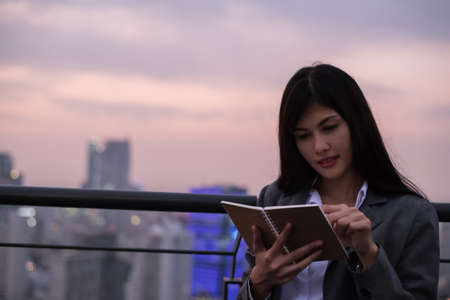 Business Concept. Asian businesswoman looking at book. Young businesswoman is happy to read. Young businesswoman reading a book on the top of a building in the evening.の写真素材