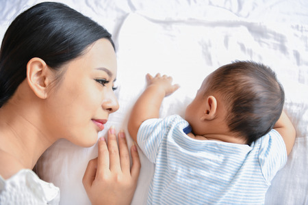 Newborn Concept. Mother and child on a white bed. Mom and baby boy playing in bedroom. Parent and little kid relaxing at home. Family having fun together. Newborn baby is fussing and crying.の写真素材