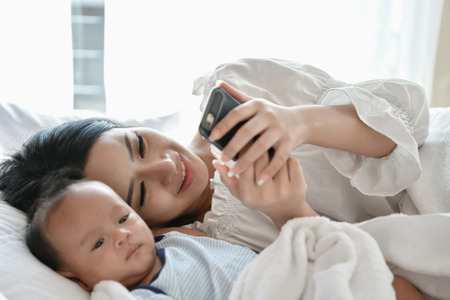 Newborn Concept. Mother and child on a white bed. Mom and baby boy playing in bedroom. Mother is...